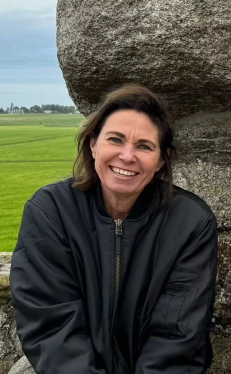 Een vrouw met schouderlang bruin haar, draagt een donker jasje en glimlacht terwijl ze buiten zit voor een stenen gebouw met groene velden en een bewolkte lucht op de achtergrond.