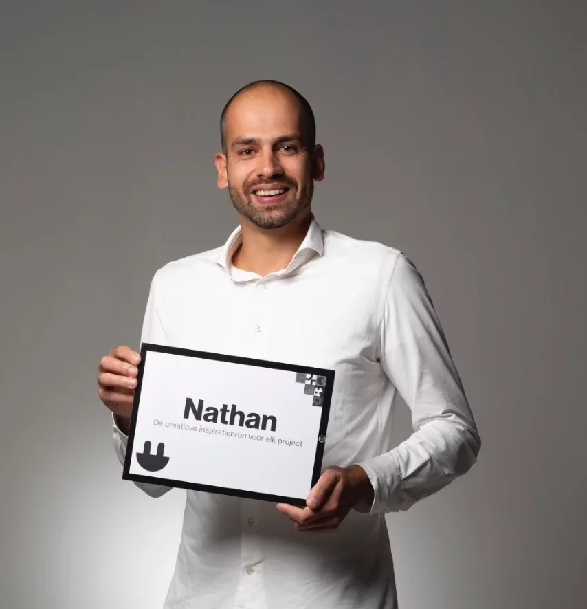 Een glimlachende man in een wit overhemd houdt een bordje vast met daarop de tekst ‘Nathan’ en daaronder wat kleinere tekst, tegen een effen grijze achtergrond.
