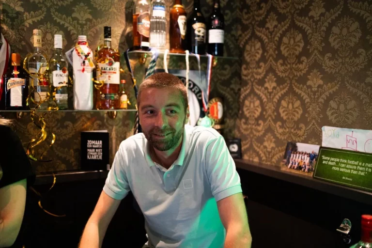 Joris, een man met kort haar en een baard die een witte polo draagt, zit aan een bar met flessen alcohol achter hem. Aan de muur hangt behang met patronen en op de toonbank hangen decoraties en foto's.