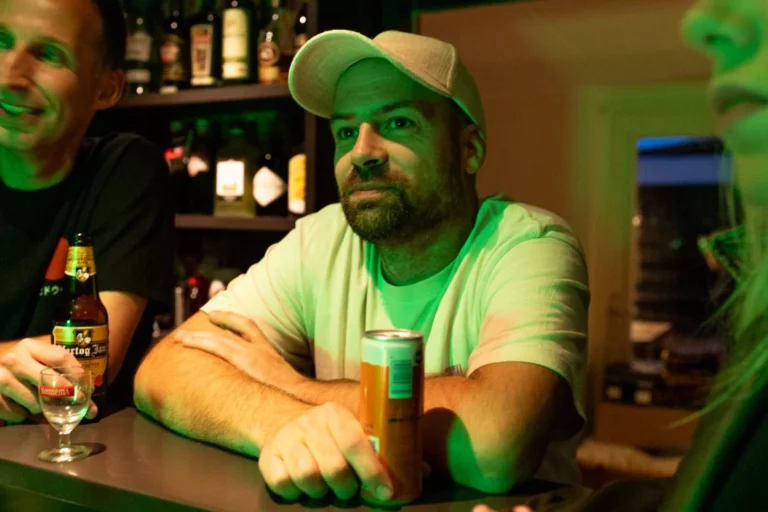 Een man in een wit T-shirt en een lichtgekleurde pet zit aan een bar met zijn armen over elkaar en houdt een oranje blikje vast. Achter hem zijn rekken met flessen zichtbaar en groene verlichting vult de scène. Andere mensen zijn gedeeltelijk zichtbaar in de buurt.