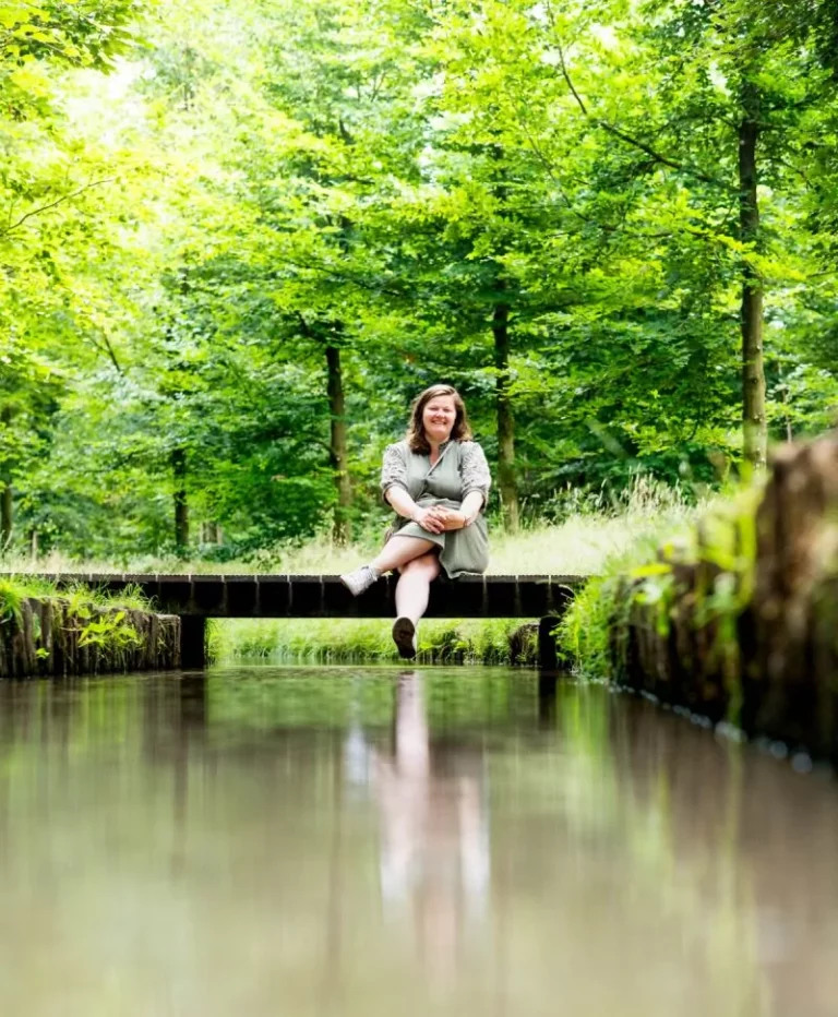 Tineke zit op een kleine houten brug over een kalm beekje, omringd door weelderig groene bomen en gras op een heldere dag.